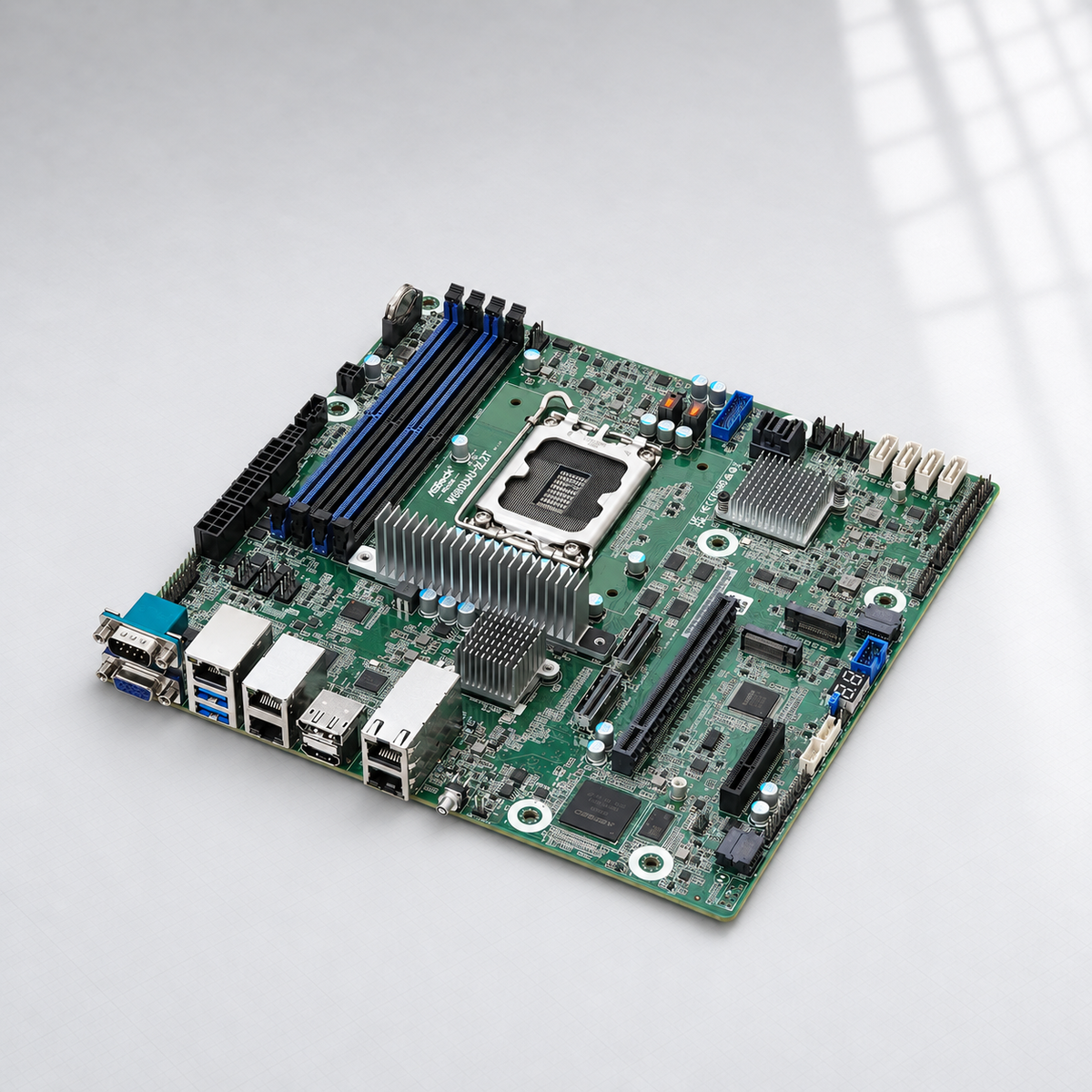 A green industrial motherboard with a CPU socket, dual blue RAM slots, multiple silver heatsinks, and various ports on the edge, photographed on a light gray background with soft window shadow overlays.
